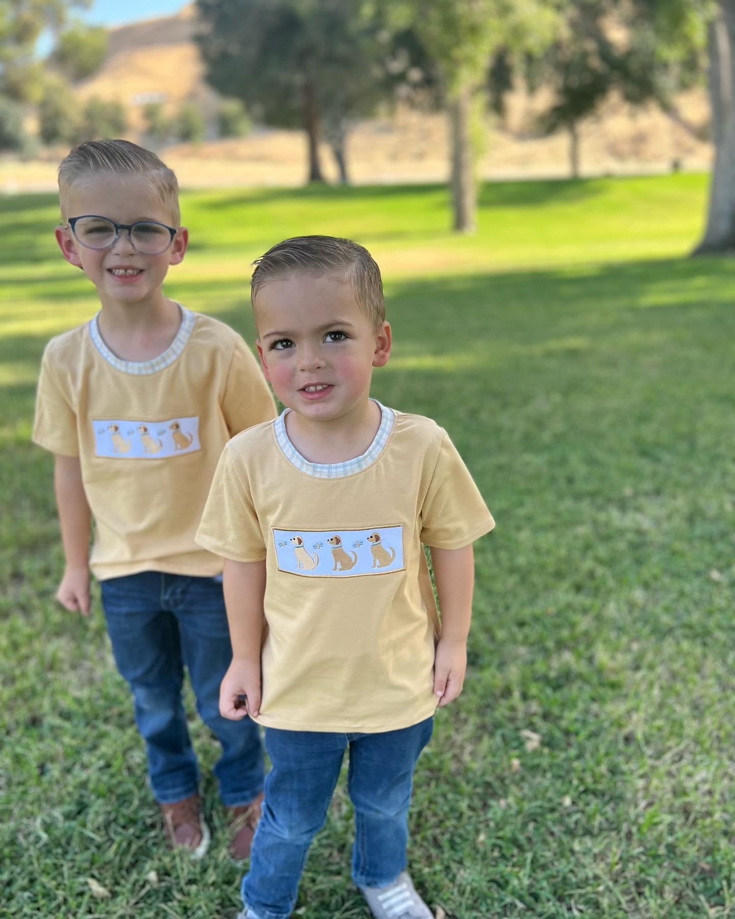 2 boys standing in a grassy park with a yellow shirt with an appliqué that has three dogs. 